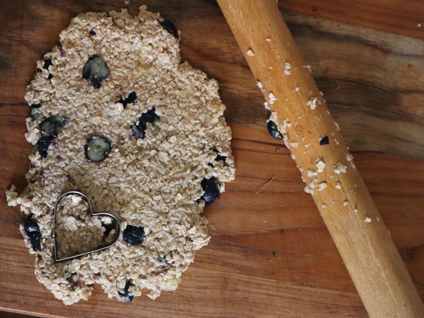 an image of an oat and and blueberry biscuit dough rolled out on a wooden chopping board with a heart-shaped cookie cutter on top, and a rolling pin laying next to it
